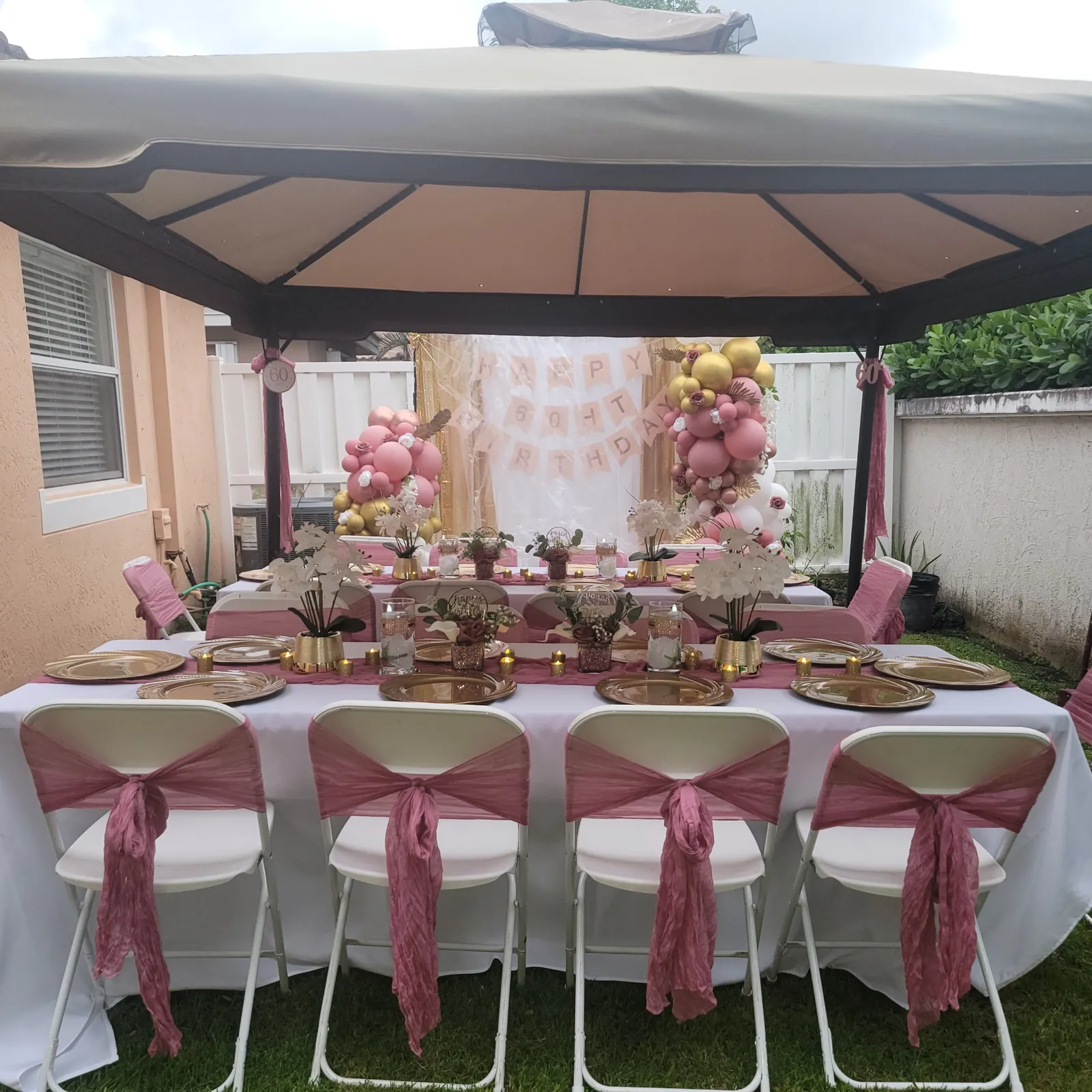 Backyard 60th birthday setup with dusty rose sashes, gold chargers, orchid centerpieces and a blush balloon backdrop