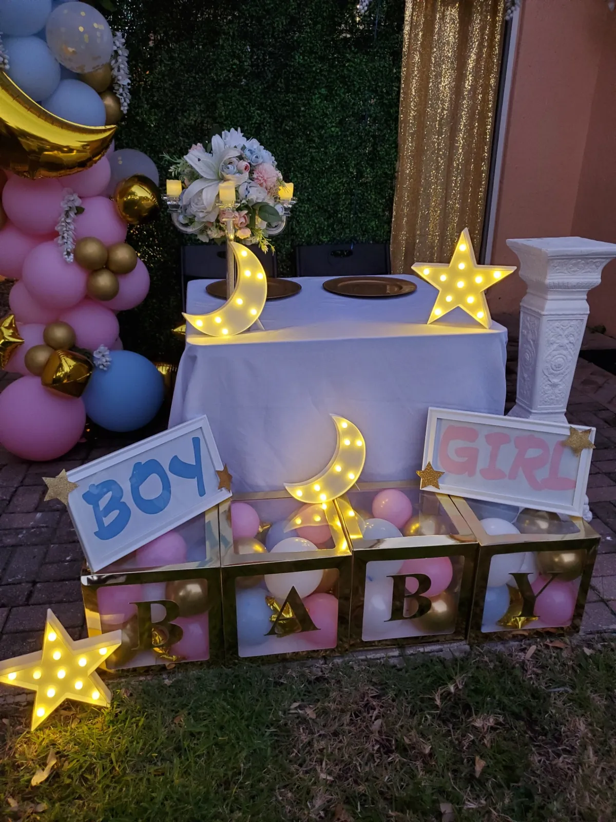 Evening gender reveal setup with pink and blue balloon arch, lighted moon and star marquees and gold BABY blocks