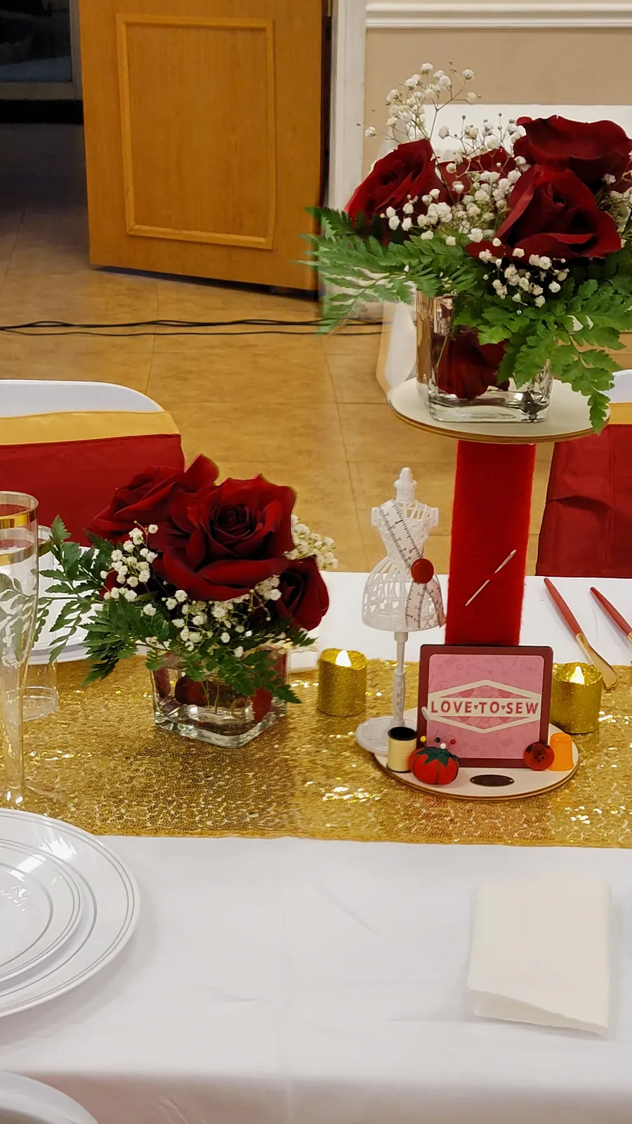 Guest table detail with red roses, gold sequin runner, mini dress form and 'Love to Sew' framed accent