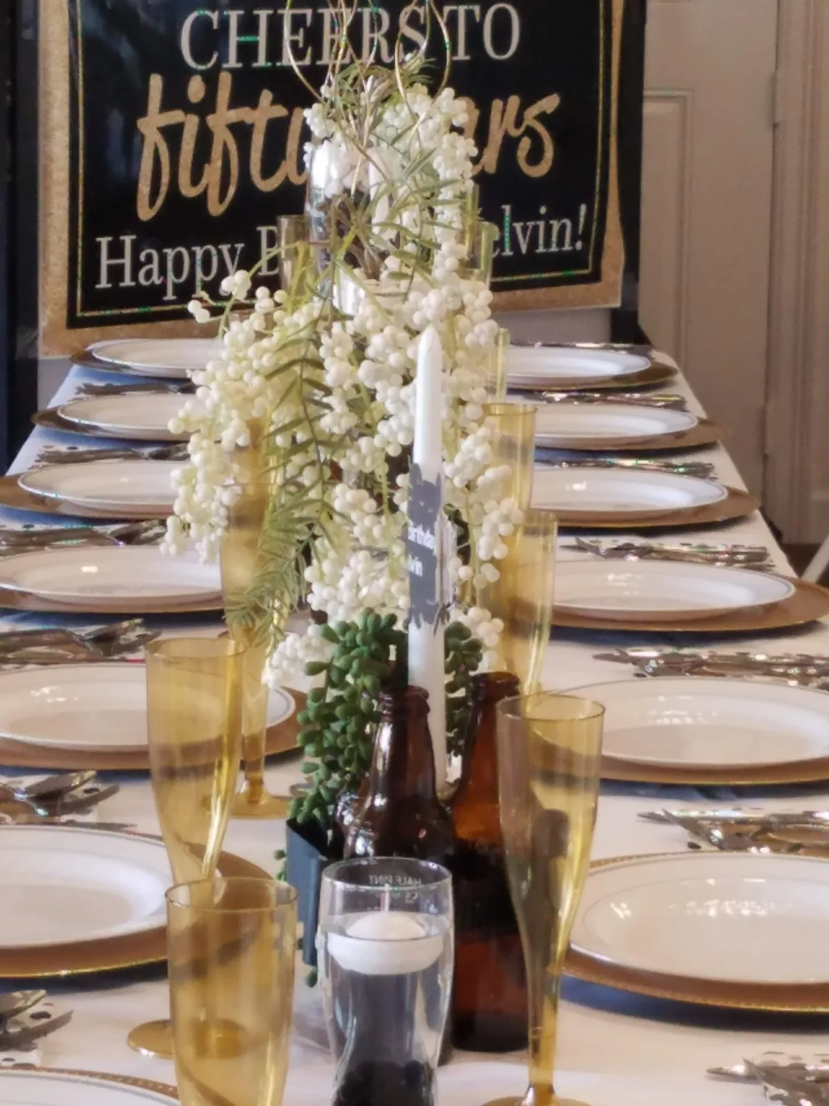 Long banquet tablescape with gold chargers, taper candles and white berry centerpieces for a milestone birthday